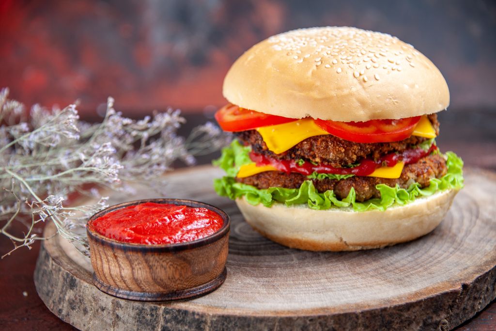 Double patty veggie burger with lettuce, cheddar cheese, tomato slices, and ketchup served with a side of tomato sauce in a wooden bowl